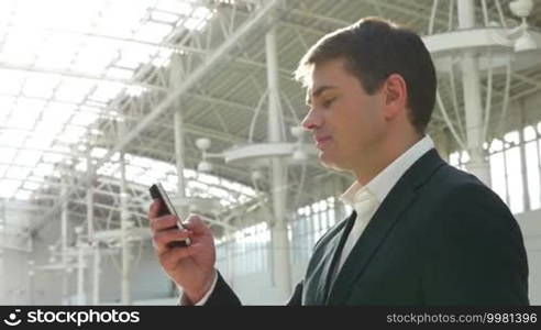 Young businessman texting on smartphone with a slight smile on the face. Metal constructions of office building or trade center in the background