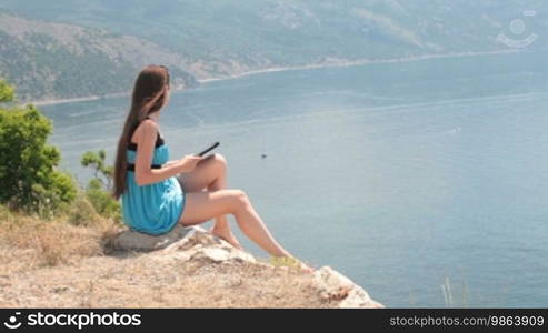 Young Caucasian woman reads an ebook on a digital e-reader device on top of a mountain by the sea