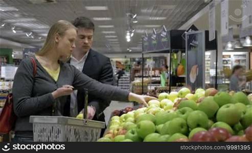 Young couple in the supermarket choosing and putting fresh apples into the shop basket. They are examining and smelling the fruit to find the best