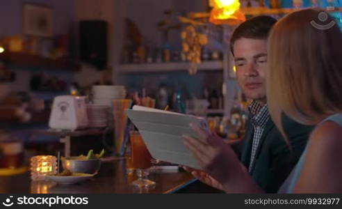 Young couple is sitting by the bar counter with tablet and drinking beverages from the glasses.