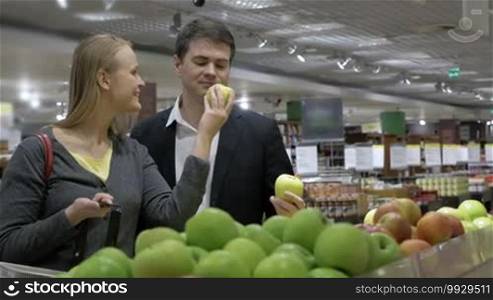 Young couple is standing in front of the stall with fruit in grocery store, choosing apples and putting them into basket.