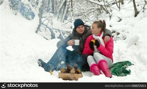 Young couple sitting by bonfire and drinking coffee in winter forest