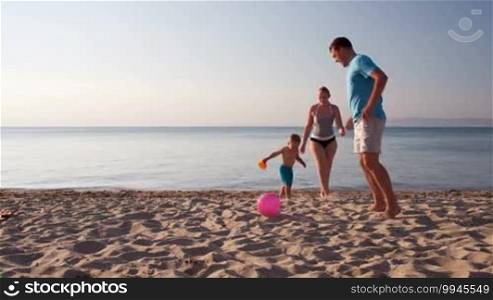 Young family frolicking together playing football soccer on the beach at sunset with the little boy running with his mother and father