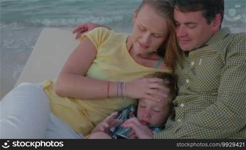 Young family relaxing on the chaise longue on the seashore. Father and son playing on cell phone while mother stroking boy's head with distracted look