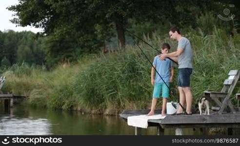 Young father and his teenage son spending leisure together fishing at freshwater pond. Handsome dad teaching boy how to fish with fishing rod and reel while standing on wooden pontoon on the lake during summer vacation.