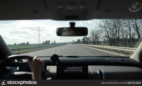 Young female driver controlling modern car on highway during summer road trip in countryside. View inside out of vehicle's interior. Charming woman driving car on freeway on sunny day.