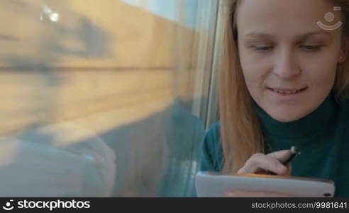 Young female passenger in the train typing SMS on smartphone with a pen. After taking it away she enjoying view from the window