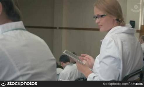 Young female student turning to the camera and smiling, then working with tablet computer during medical conference or lecture