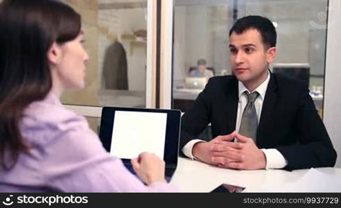 Young handsome man in corporate clothing sitting at the desk and talking about his successful work experience during a job interview at the office. Young brunette female headhunter interviewing a candidate to fill a business position.