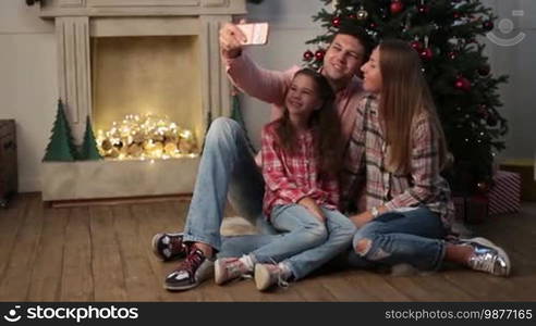 Young happy family of three sitting on the floor by the Christmas tree and taking a selfie on a smartphone in a cozy dark decorated room. Joyful parents and a cute daughter making a selfie on a mobile phone during winter holidays. Dolly shot.