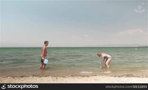 Young happy man and woman having fun at the seaside. They are playing tennis with paddles standing in the sea. The man is falling into the water trying to get the ball