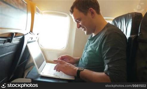 Young happy man using laptop in the airplane. He typing message and looking out illuminator while waiting for an answer. Comfortable and fast traveling