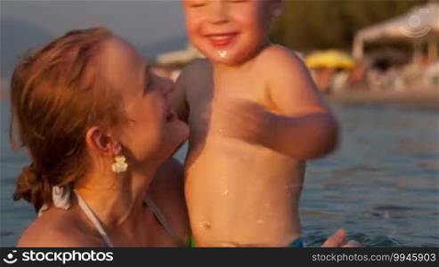 Young happy mother toss up her little three-year-old sonny while swimming in the sea near the beach, smiling and laughing while jumping