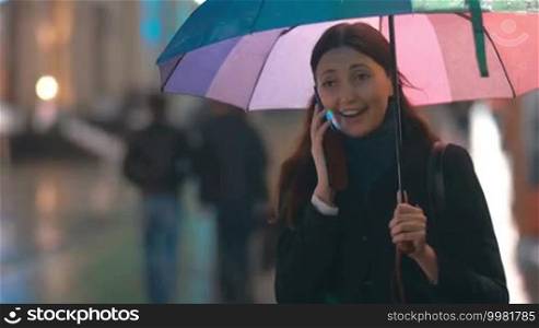 Young happy woman having exciting phone talk holding colorful umbrella in rainy evening in the city