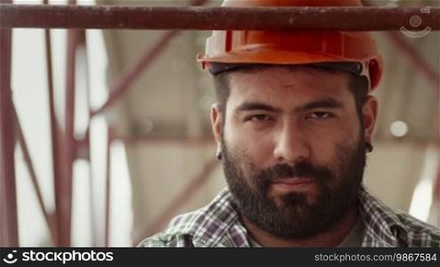 Young Hispanic man at work in construction site, portrait of serious manual worker with helmet looking at camera. Part 2 of 11