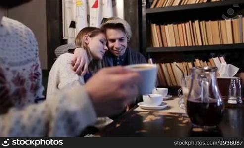 Young lovers hugging, caressing, chatting and laughing while sitting in coffee shop together with their friends. Foreground male teenager holding cup of coffee and drinking. Pretty teenage couple in love hugging and expressing their feelings in cafe.