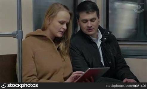 Young man and woman in a moving underground train. They are using a tablet and talking during the ride