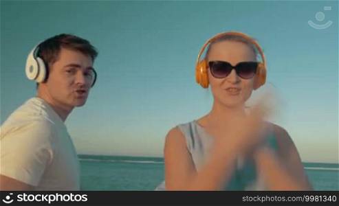 Young man and woman relaxing with favorite music on the beach. They are wearing wireless earphones, singing, and dancing