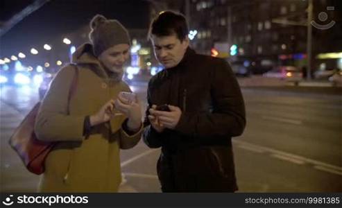 Young man and woman standing by the busy road in the evening city and explaining something to each other using cellphones