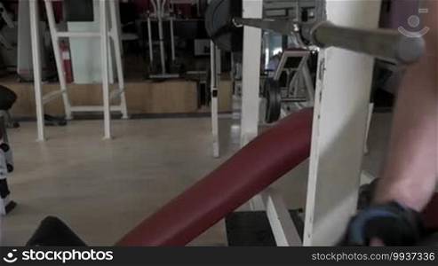 Young man at the gym, hangs up dumbbell on rod and then lies on bench to do chest press exercise with rod