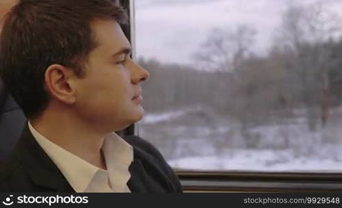 Young man in business suit is sitting in train carriage. He's looking out the window and thinking.