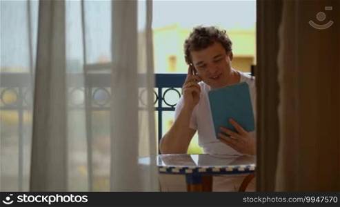 Young man talking on the phone with a tablet PC in hands on a hotel balcony