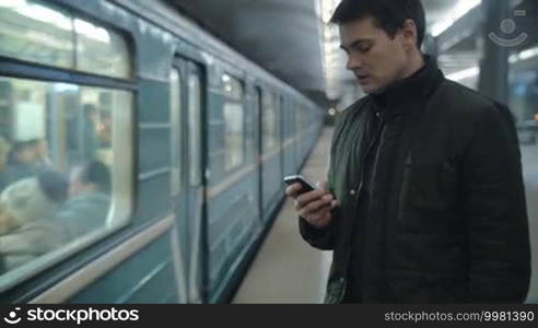 Young man using smartphone to type text message at the underground station, train passing by. Everyday traveling by public transport