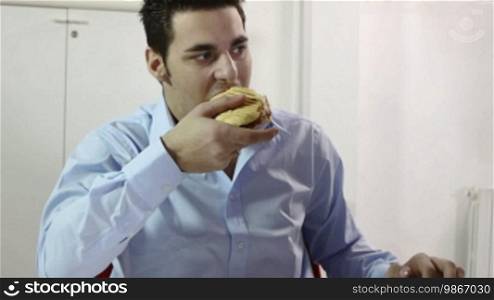 Young manager at work, eating hamburger and working with computer