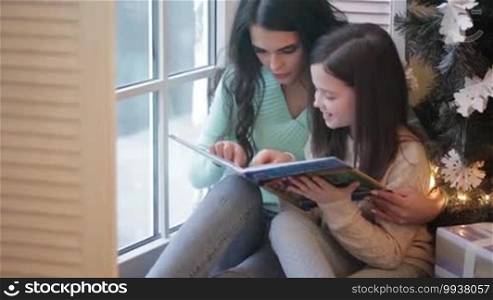 Young mother and daughter reading book by the window