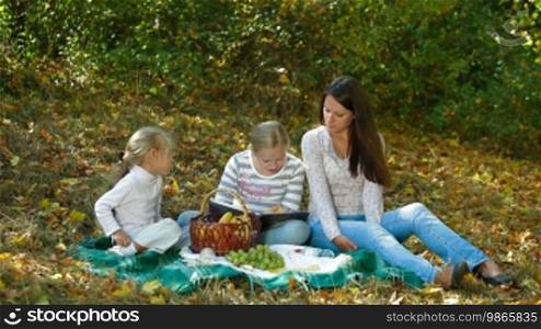 Young mother and two daughters enjoying an autumn day at a picnic in the park, child reading a book