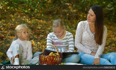 Young mother and two daughters enjoying autumn day at picnic in the park, child reading a book