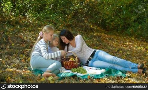 Young mother and two daughters having a picnic in the autumn park, talking and eating fruit