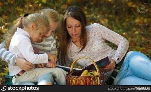 Young mother and two daughters having a picnic in the autumn park, reading a book. Medium shot