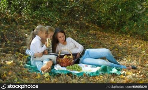 Young mother and two daughters having a picnic in the autumn park, reading a book. Long shot