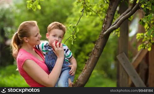 Young mother feeding her son with a red fresh apple in the orchard. Healthy food