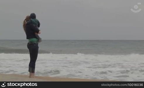 Young mother with her little son staying on the beach and looking at the sea waves. No faces visible.