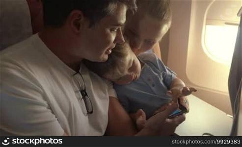 Young parents and little son traveling by air. Child playing game on father's smartphone to entertain himself during the flight. Mother kissing the child