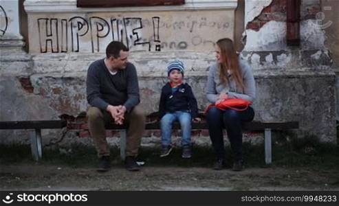 Young parents and their little son sitting on the bench near a worn grungy building with a Hippie inscription on the wall