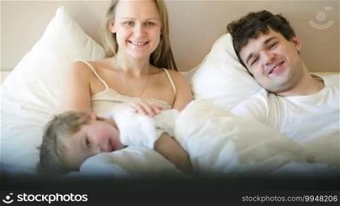 Young parents and their son watching TV lying in the bed in the morning. Father taking remote control and changing channel