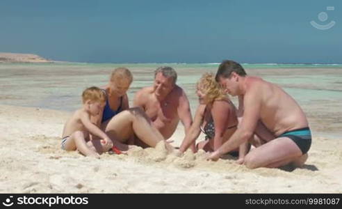 Young parents, little son, and grandparents on summer vacation. They are sitting on the beach by the sea and building a sandcastle