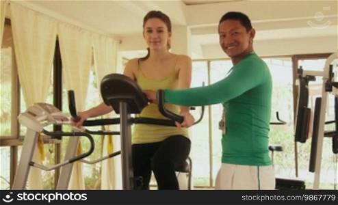 Young people and leisure, lifestyle and fun, travel and vacation, man and woman on holidays. Happy pretty girl exercising in fitness club with personal trainer in sport gym, smiling at camera. 3 of 12
