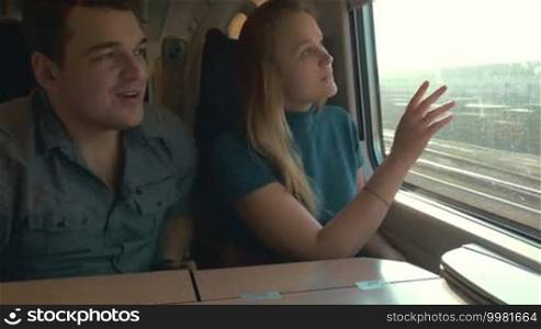 Young people traveling by train. Woman is curious about the places she sees in the window, while man is filming them with a retro video camera
