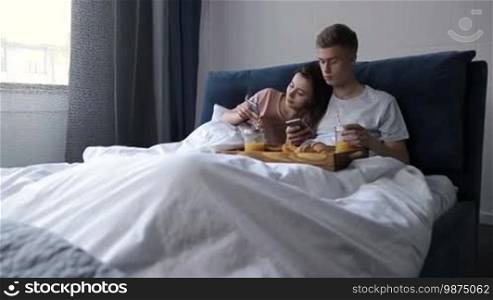 Young positive couple lying in the bed, browsing social networks on mobile phones while having breakfast in bedroom in the morning. Hipster family enjoying time together, communicating and using mobile phones in comfortable bed at home.