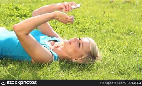 Young pretty blonde girl lying on her back in the green grass reading text messages on her mobile phone