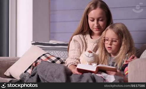 Young pretty mother and her little daughter wrapped in a blanket sitting on the sofa in a nice cozy room and reading bedtime story together. Sweet child learning to read with her mother at home.