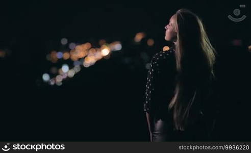 Young sensual woman is standing on the observation deck and enjoying panorama of city at night. The wind is blowing in her long nice hair. On background city streetlights.