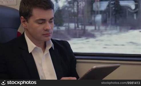 Young serious businessman traveling by train and using a tablet computer, sometimes getting distracted by the view out the window. Business trip
