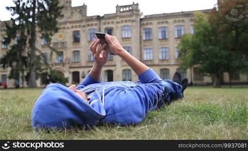 Young smiling male student in hoodie lying on the grass after classes outside university campus and browsing on the cellphone