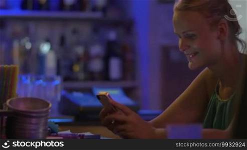Young smiling woman is sitting by the bar counter with smartphone in hands. Bartender is putting a glass with cocktail in front of her.