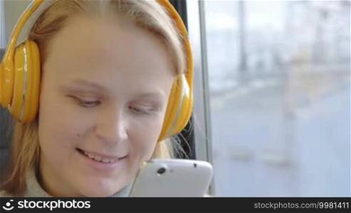 Young smiling woman traveling by train with music. She using cell phone and enjoying favorite songs in wireless headphones